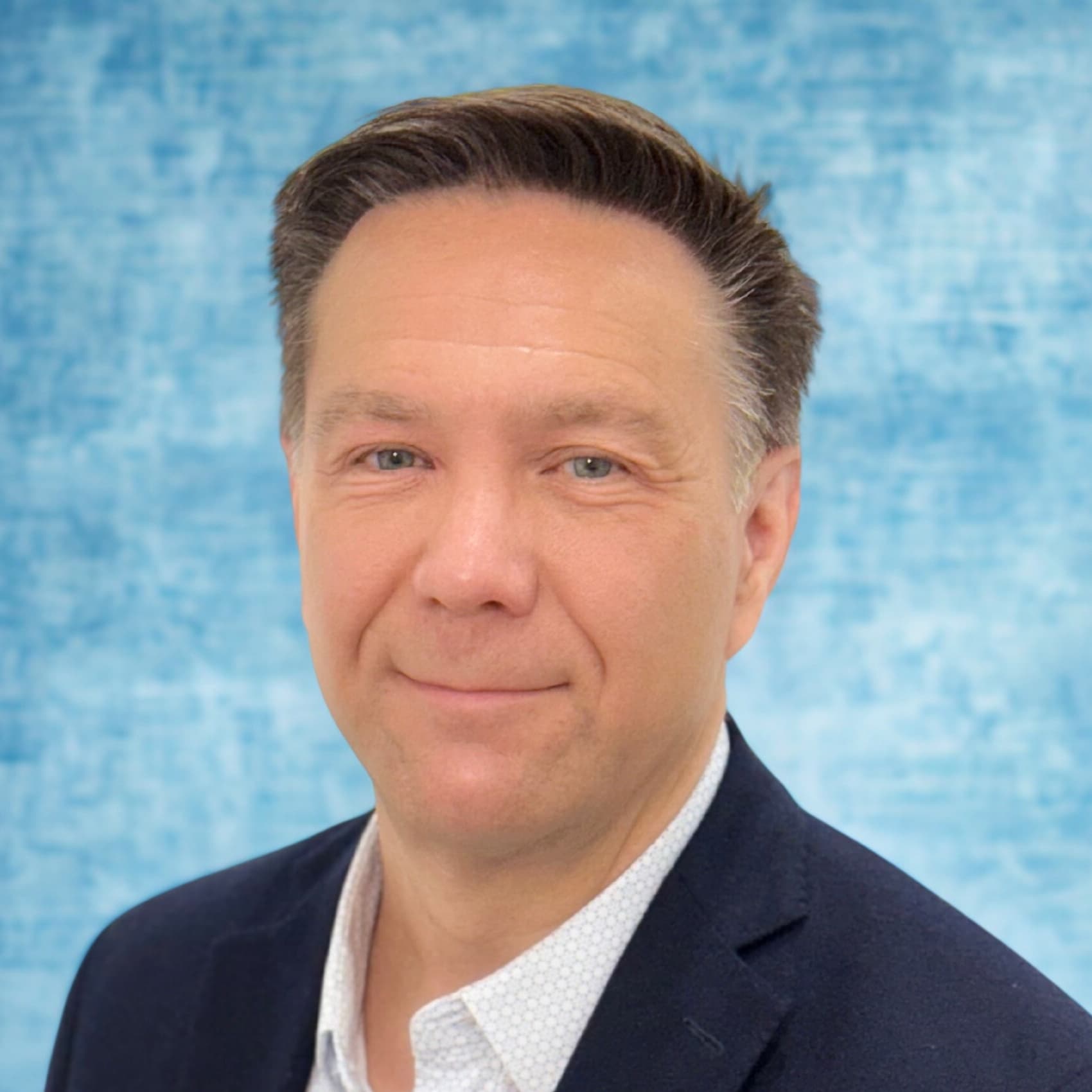 Headshot of Shawn Slusser in a navy blazer and light patterned shirt against a mottled blue studio backdrop.