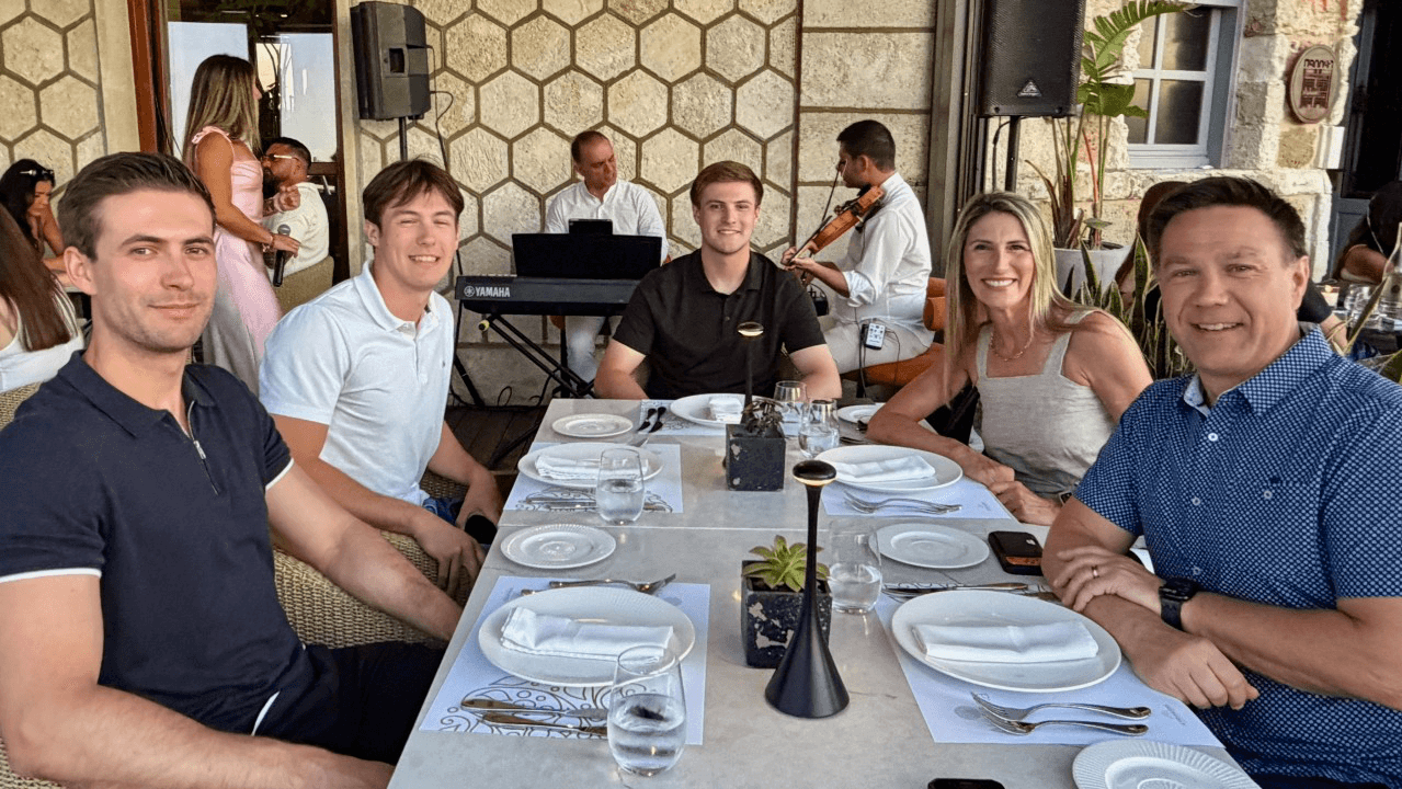 Shawn Slusser, his wife, and their three adult sons smiling around a table at an outdoor restaurant on a family vacation, with a live band playing behind them.